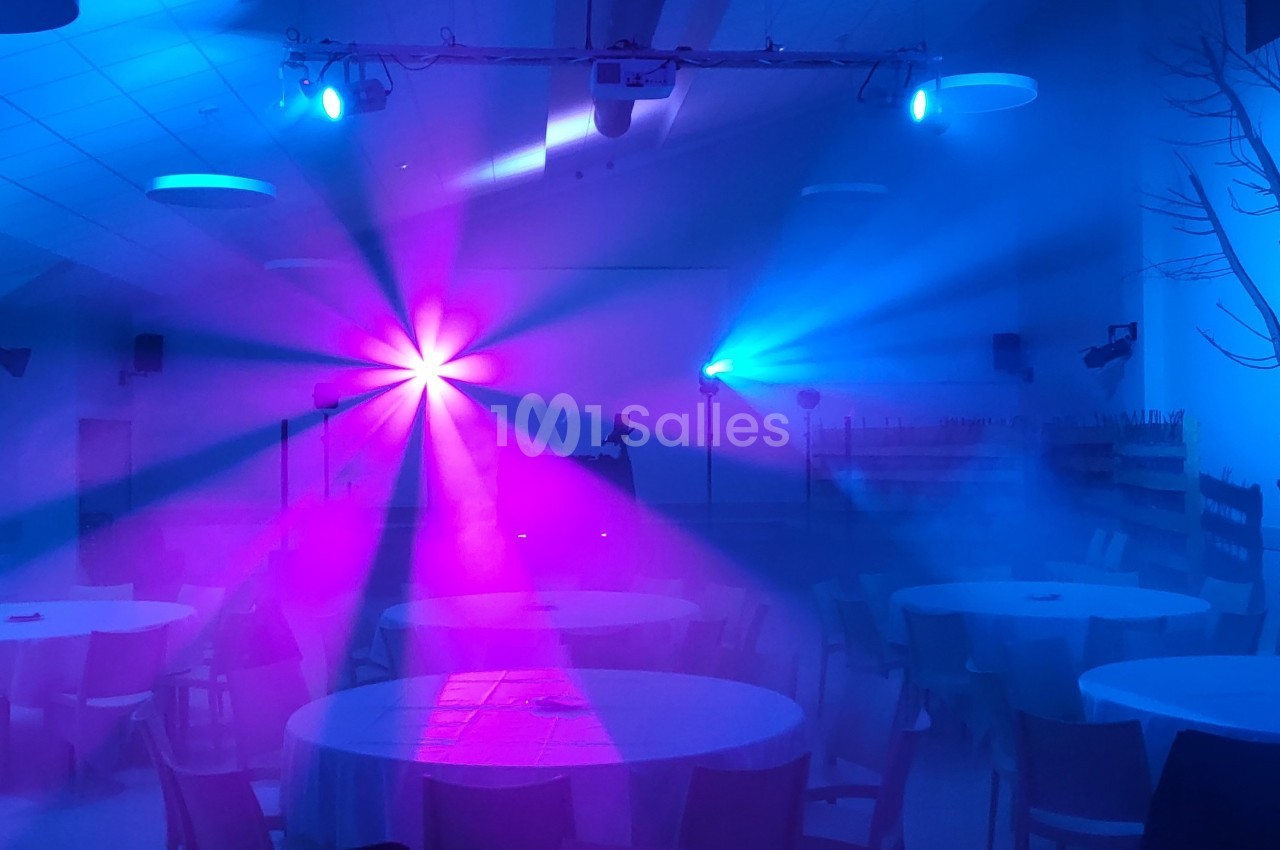 Salle décorée avec des tables rondes, éclairée par des lumières colorées bleues et roses créant une ambiance festive.