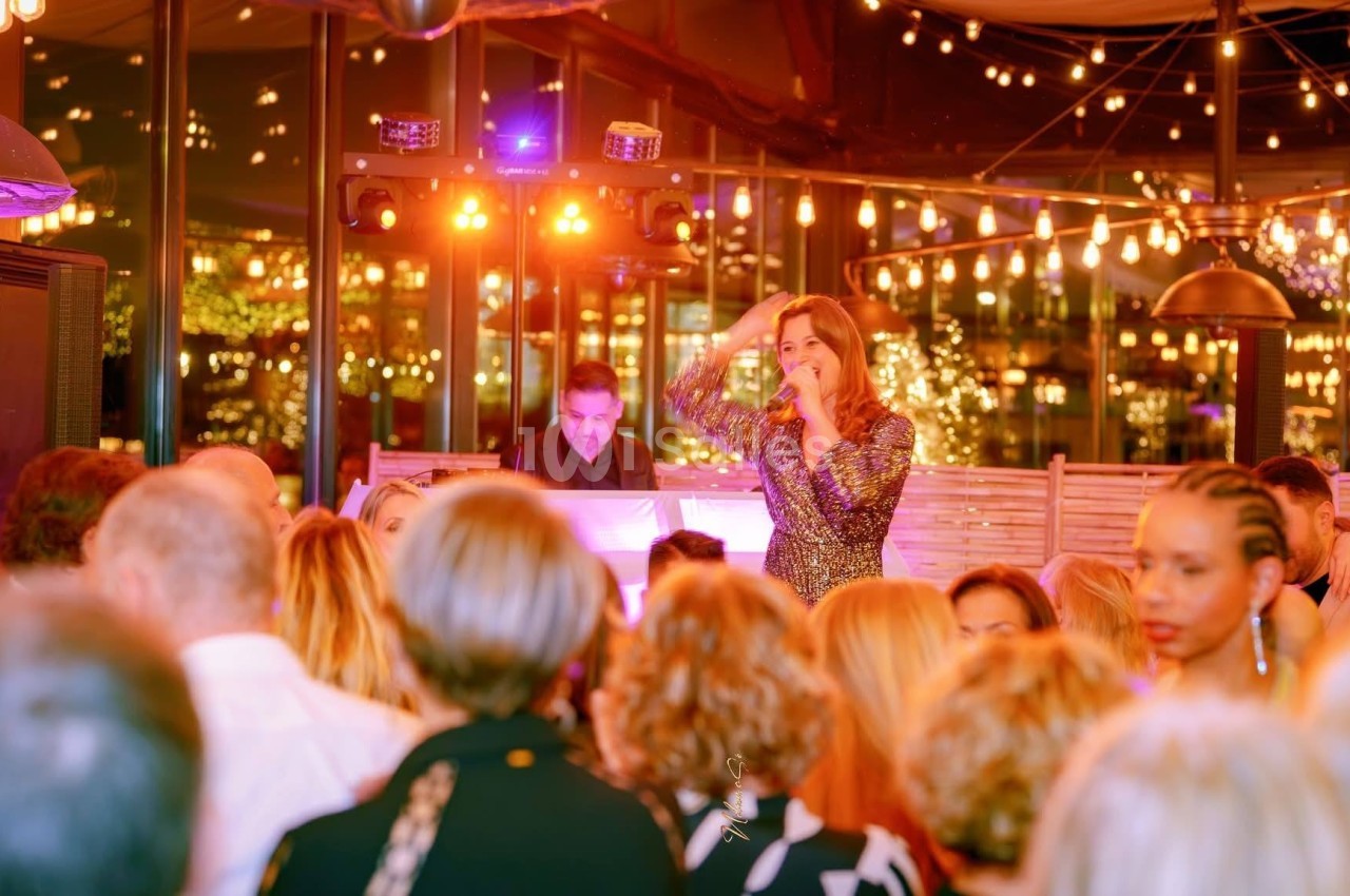 Une chanteuse en robe scintillante se produit devant un public dans une salle éclairée par des guirlandes lumineuses.