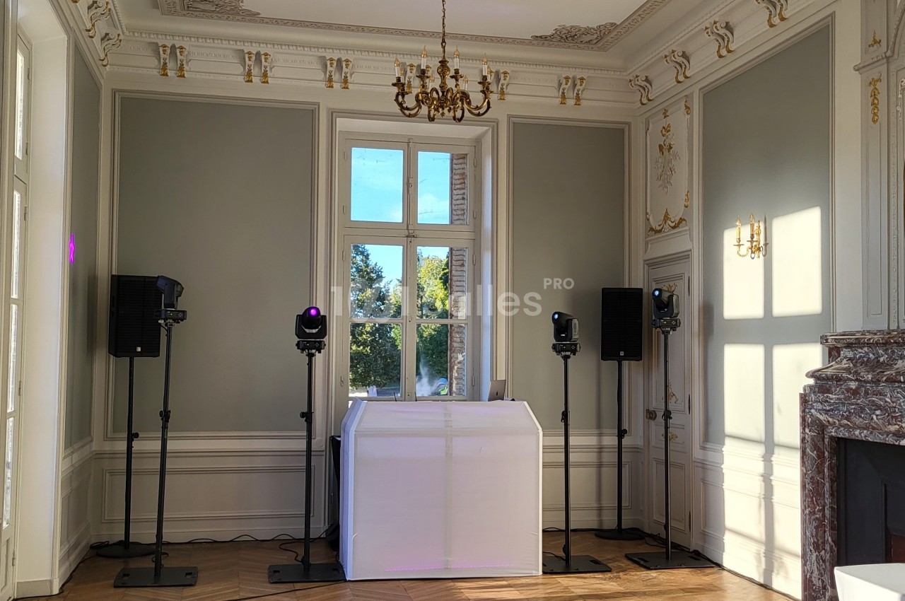 Salle élégante avec moulures, cheminée en marbre, lustre doré et installation de matériel audio devant une fenêtre.