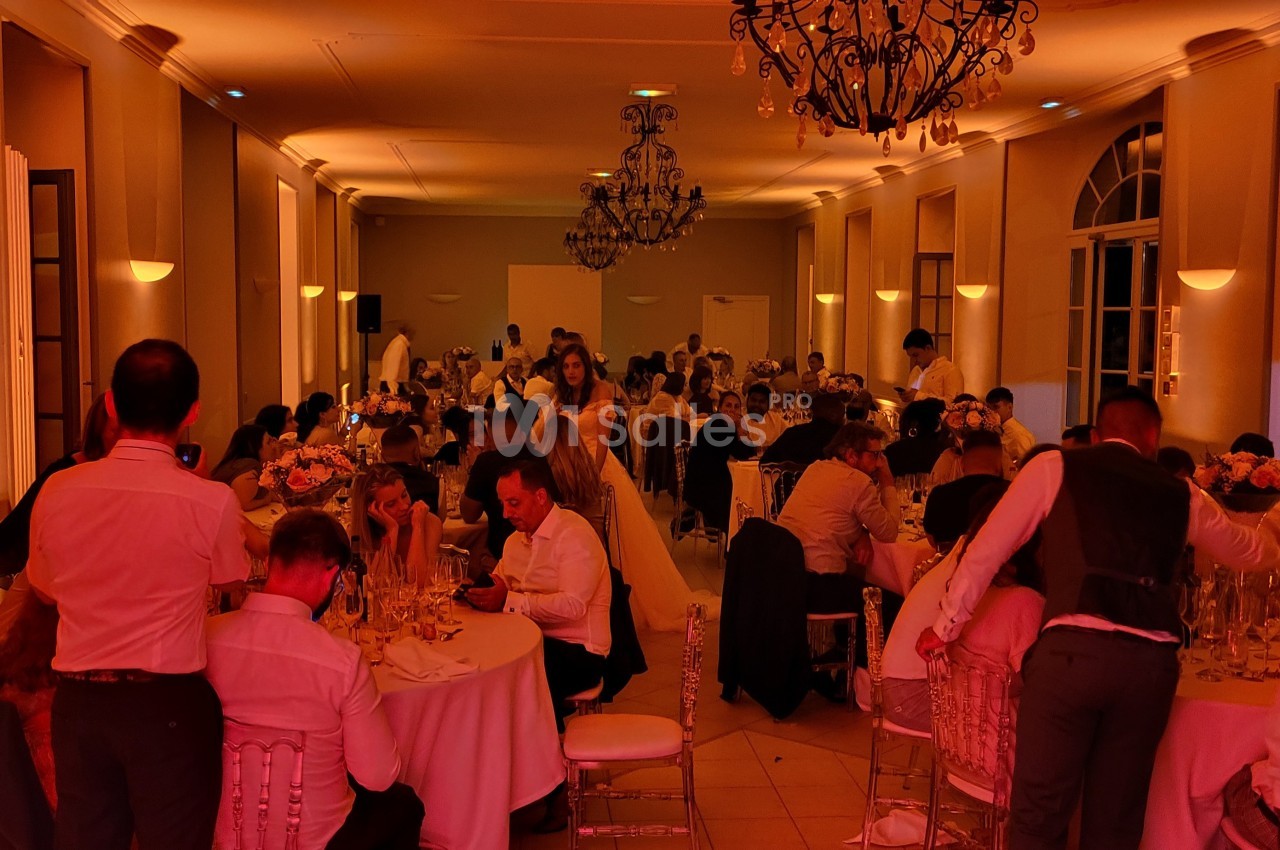 Salle de réception éclairée en rouge, avec des convives assis à des tables rondes lors d'un événement festif.