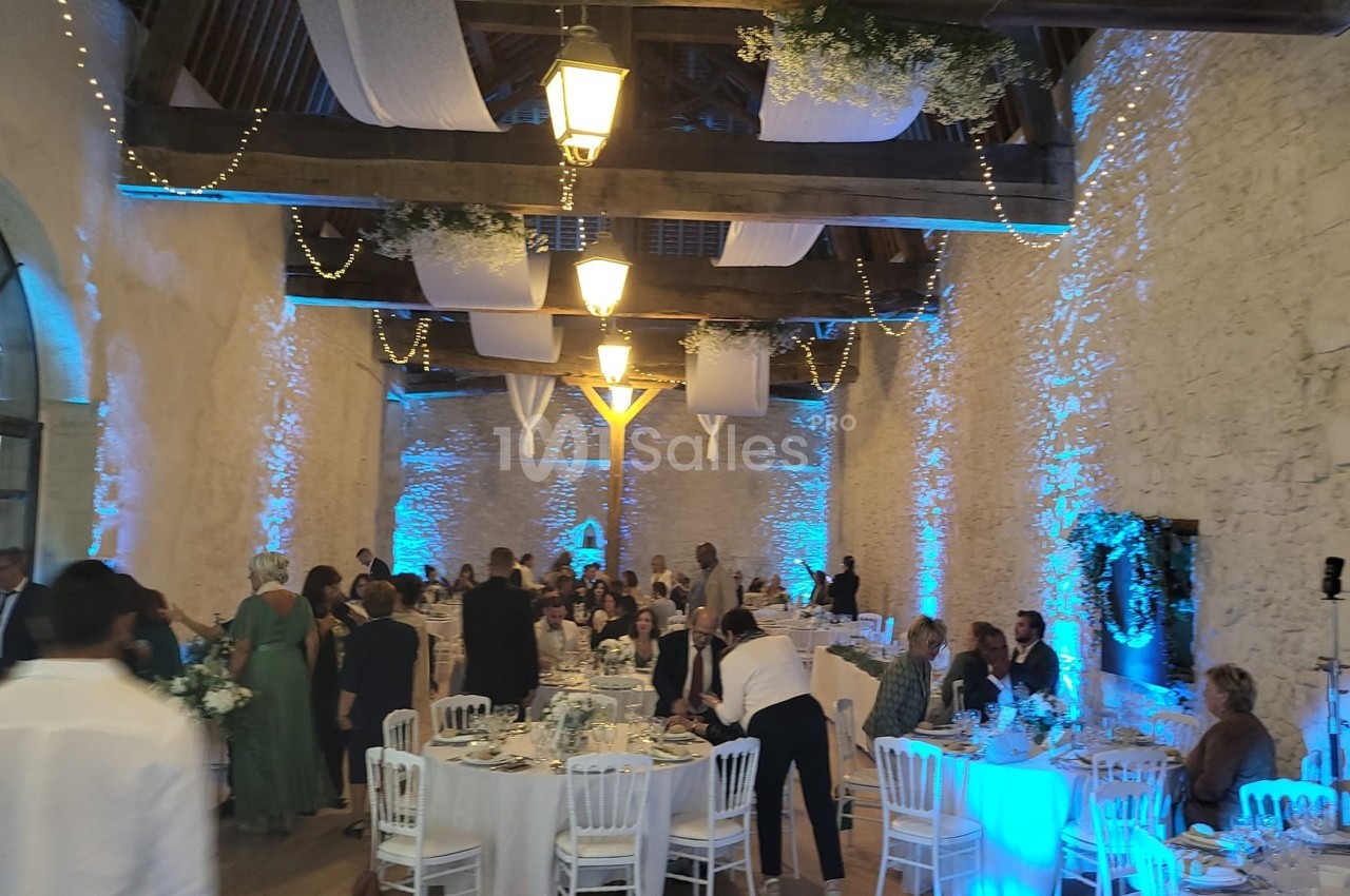 Salle de réception en pierre avec tables dressées, invités debout et éclairage bleu et guirlandes suspendues.