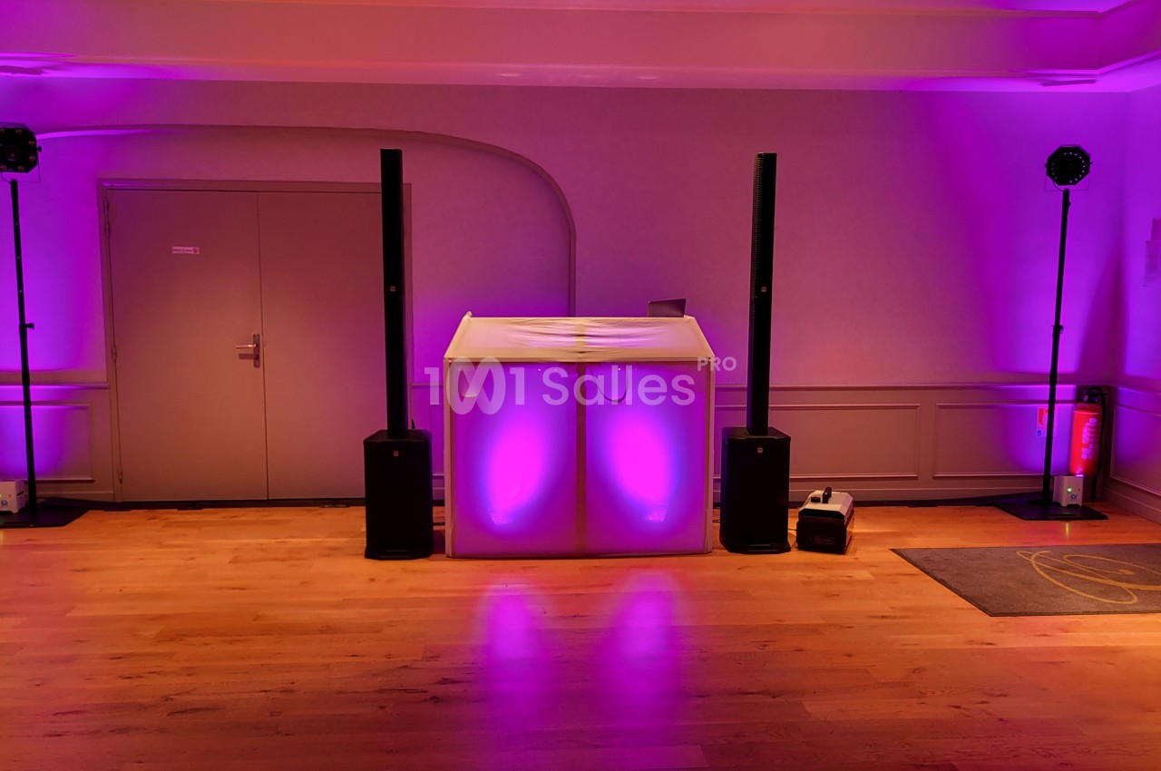 Salle éclairée en violet avec une piste en bois, une table de DJ lumineuse et des enceintes noires.