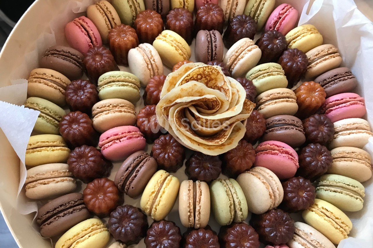 Assortiment de macarons colorés et de cannelés disposés en cercle autour d'une rose en crêpe.