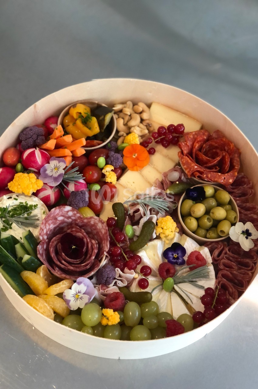 Plateau rond garni de charcuteries, fromages, fruits, légumes, olives et fleurs comestibles, disposés harmonieusement.