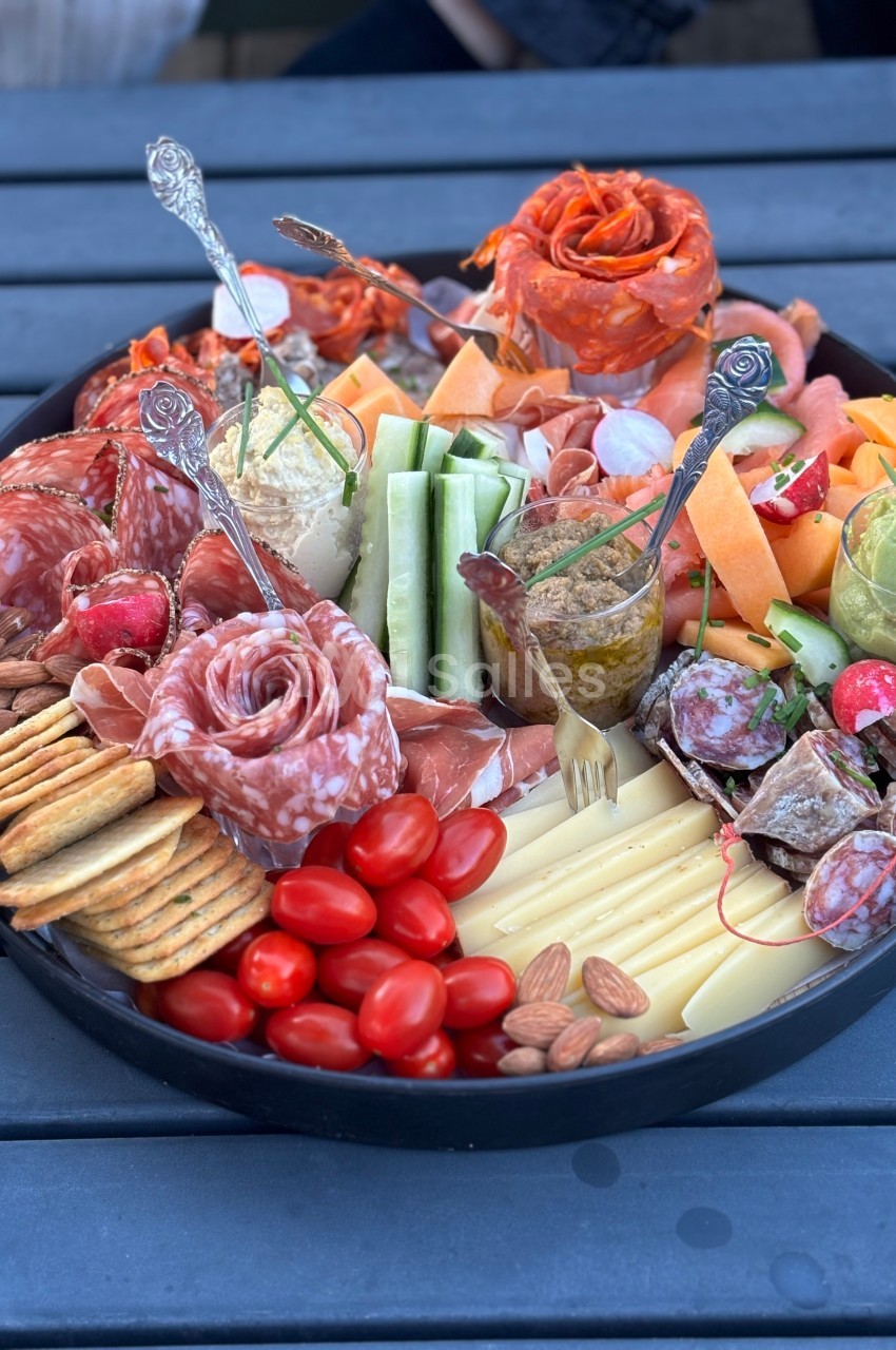 Plateau garni de charcuterie, fromages, légumes frais, fruits secs, tomates cerises et crackers sur une table en extérieur.