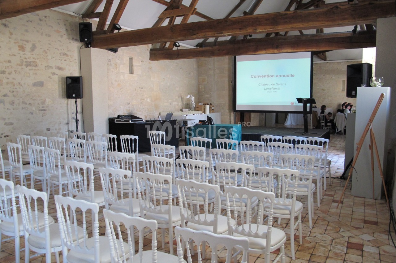 Salle de conférence avec chaises blanches alignées, écran de projection allumé et poutres apparentes au plafond.