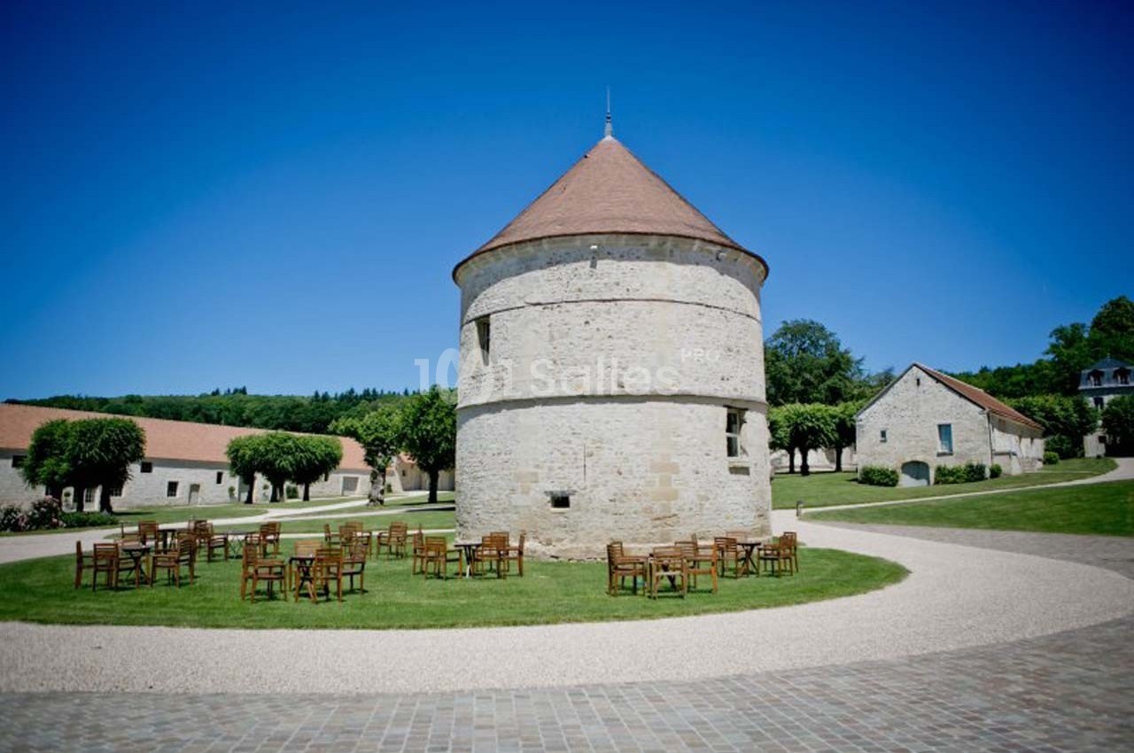 Tour en pierre ronde avec toit en tuiles, entourée de chaises en bois sur une pelouse, dans un cadre rural ensoleillé.