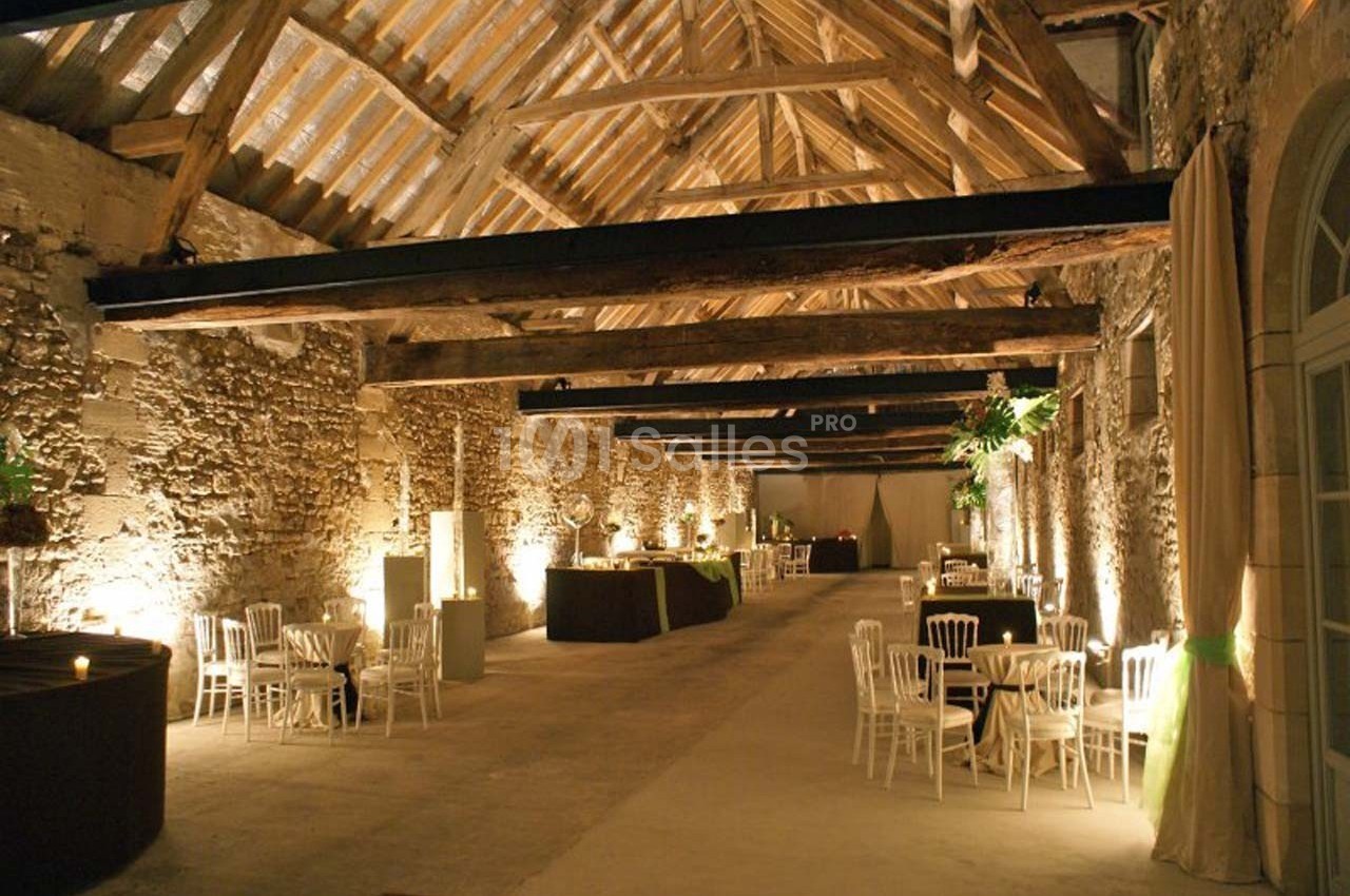 Salle en pierre avec poutres apparentes, aménagée pour un événement avec tables rondes et chaises blanches.