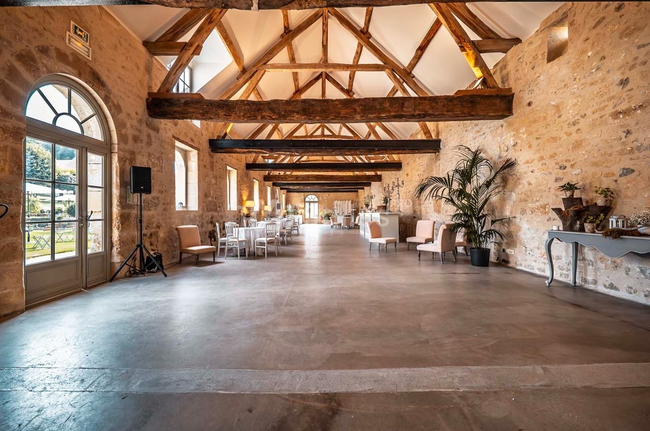 Salle spacieuse avec poutres apparentes, murs en pierre, grandes fenêtres et mobilier disposé pour un événement.