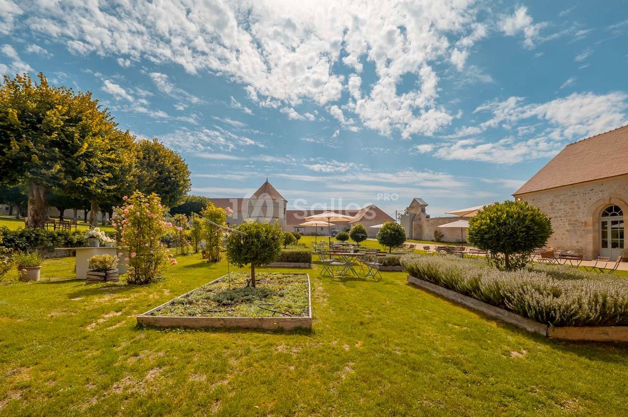 Jardin aménagé avec parterres de plantes, tables et chaises, entouré de bâtiments en pierre sous un ciel partiellement…