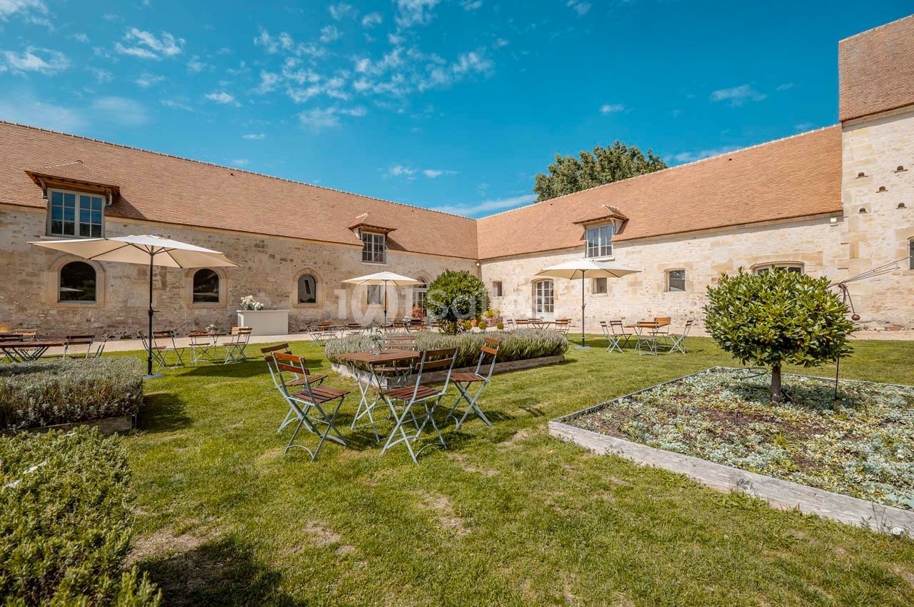 Cour intérieure avec tables et chaises en métal, parasols, pelouse et bâtiments en pierre sous un ciel bleu.