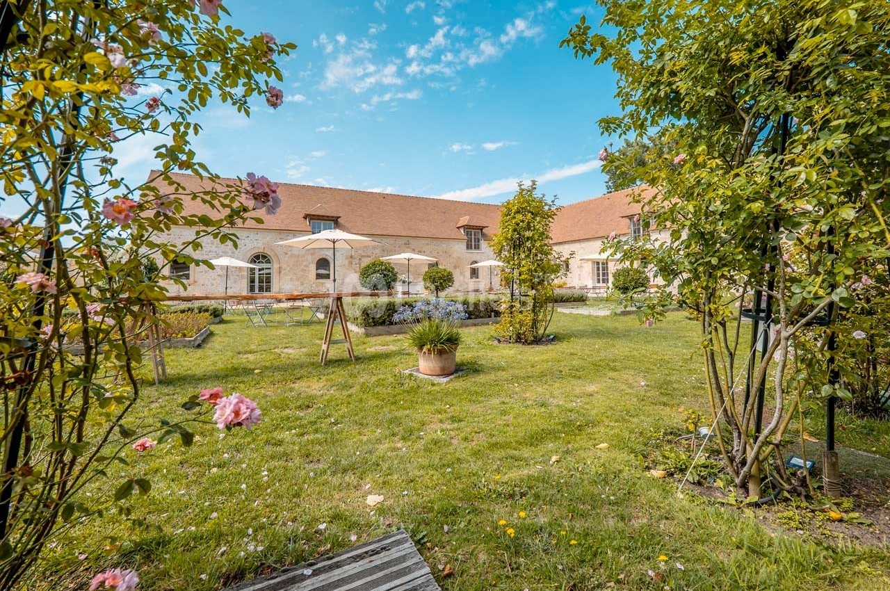 Jardin verdoyant avec fleurs, arbres et mobilier de jardin devant un bâtiment en pierre sous un ciel bleu.