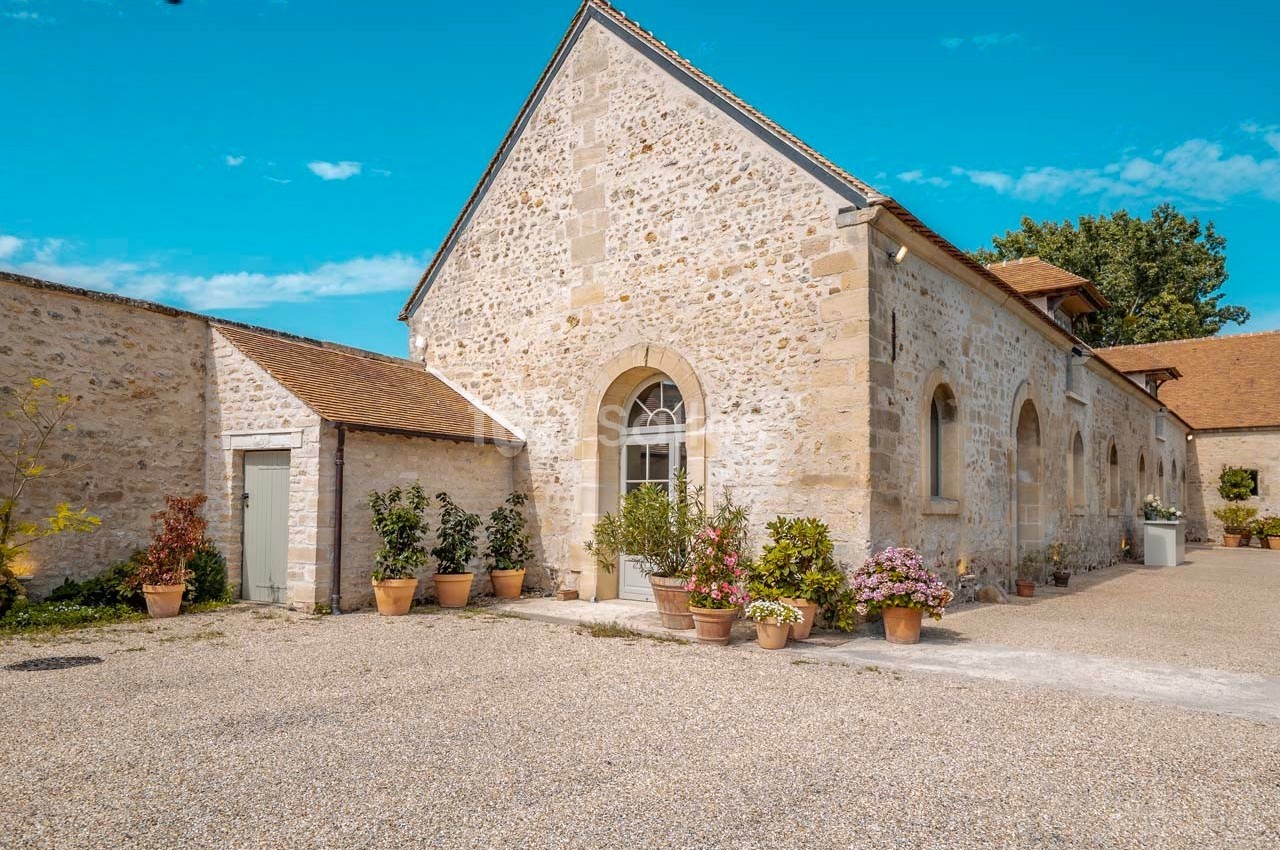 Bâtiment en pierre avec toit en tuiles, entouré de pots de fleurs, dans une cour en gravier sous un ciel bleu.