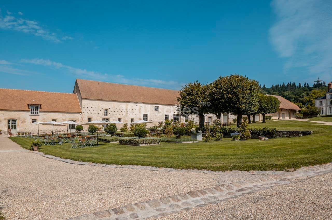 Bâtiment en pierre avec toit en tuiles, entouré d'un jardin aménagé et d'une pelouse sous un ciel bleu.