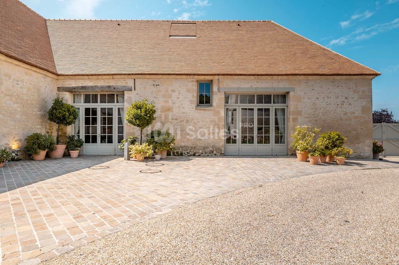 Façade en pierre d'un bâtiment avec toit en tuiles, grandes portes vitrées et plantes en pots sur une cour pavée.