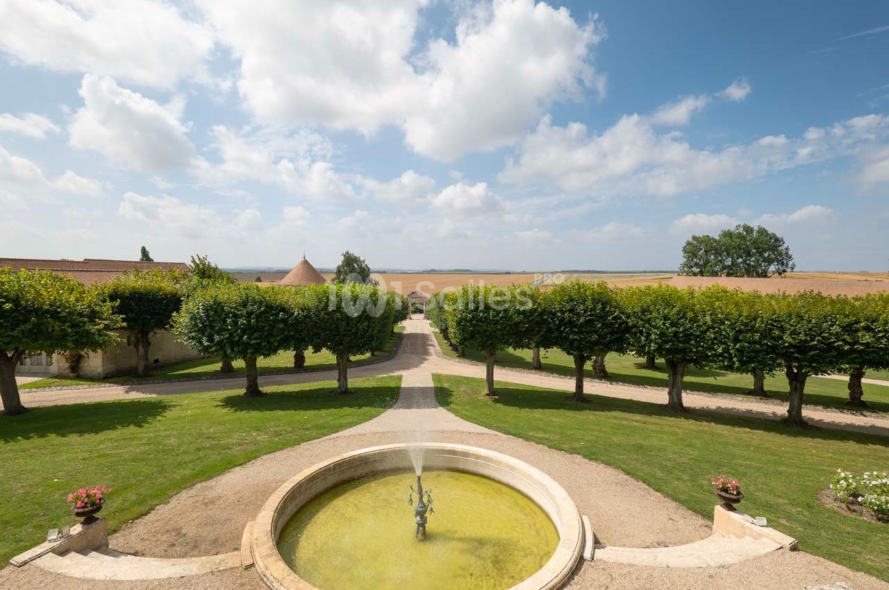 Vue d'une allée bordée d'arbres menant à des bâtiments, avec une fontaine circulaire au premier plan et un champ à l'arrière.