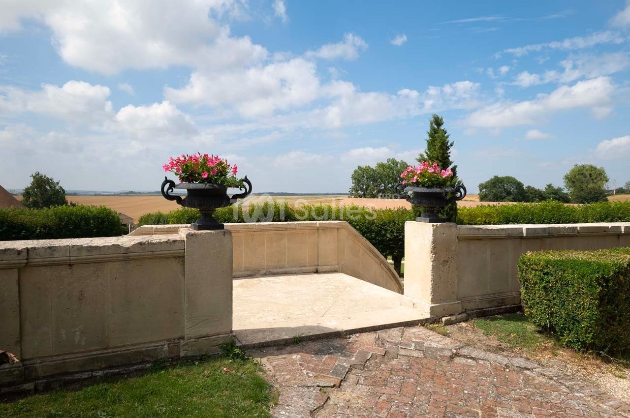 Vue sur une terrasse en pierre avec deux jardinières fleuries, donnant sur un paysage de campagne sous un ciel bleu.