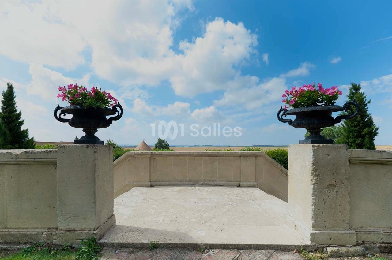 Deux jardinières fleuries sur des piliers encadrent une terrasse offrant une vue dégagée sur la campagne sous un ciel bleu.