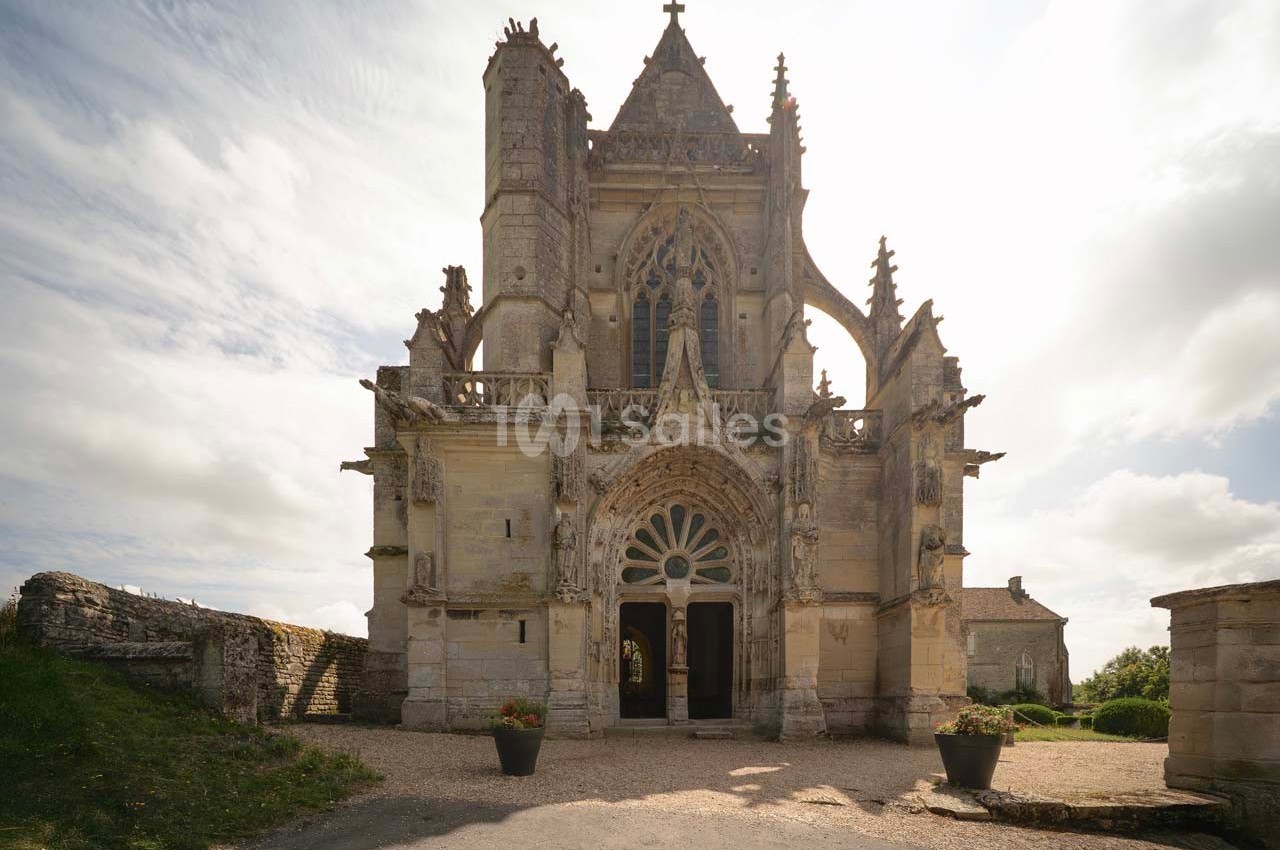 Façade d'une petite église gothique avec des détails sculptés, entourée de végétation et éclairée par une lumière douce.
