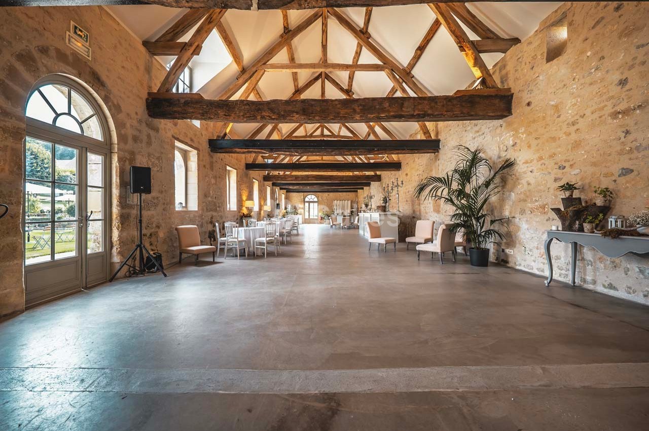 Salle lumineuse avec poutres apparentes, murs en pierre, grandes fenêtres et mobilier disposé pour un événement.