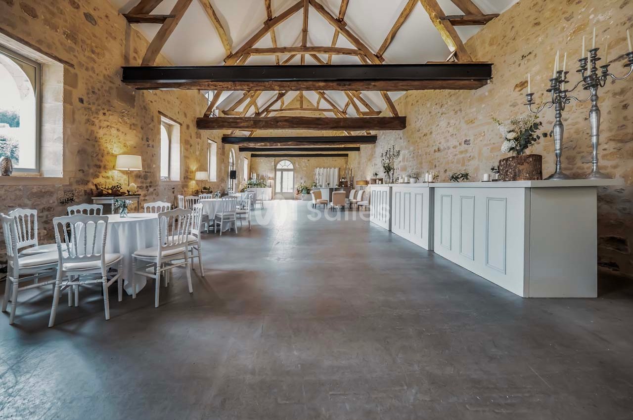 Salle lumineuse avec murs en pierre, poutres apparentes, tables rondes dressées et un comptoir blanc sur la droite.
