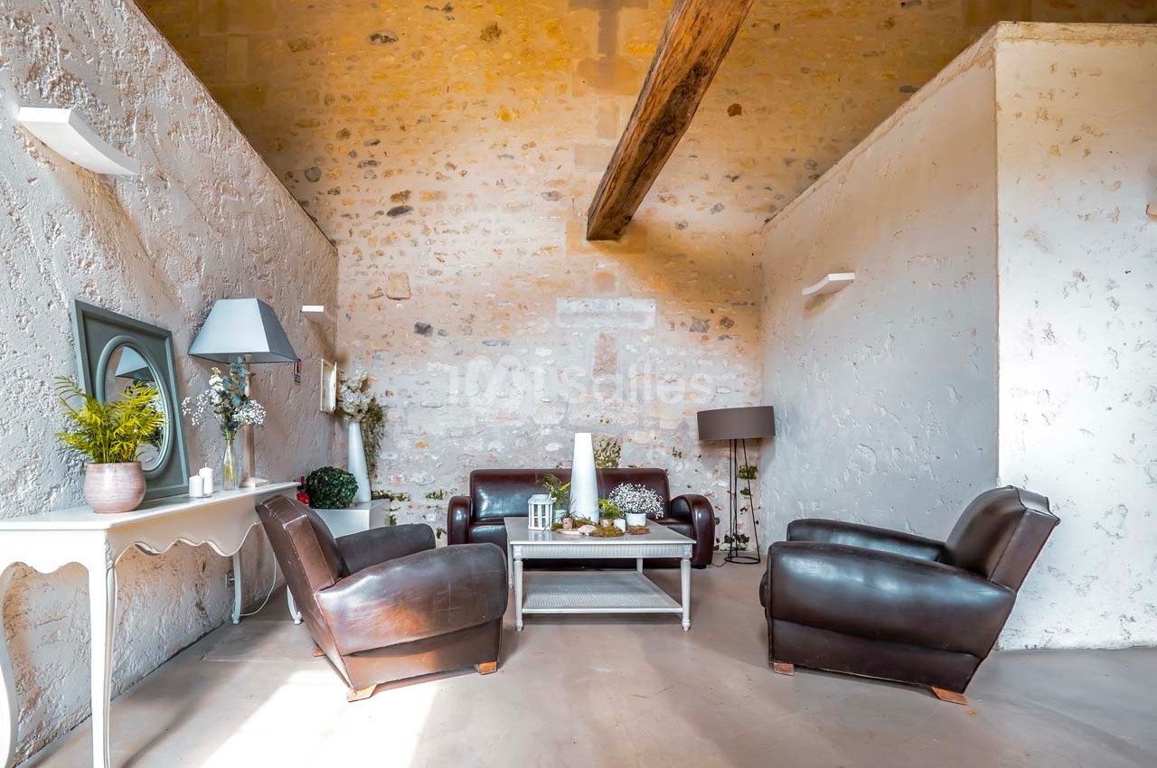 Salon lumineux avec murs en pierre, poutre apparente, fauteuils en cuir marron et table basse blanche.