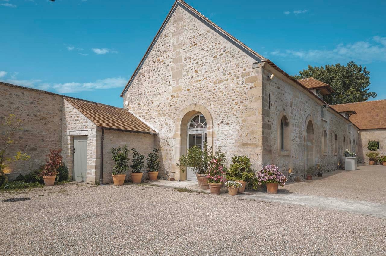 Bâtiment en pierre avec toit en tuiles, entouré de pots de fleurs, dans une cour en gravier sous un ciel bleu.