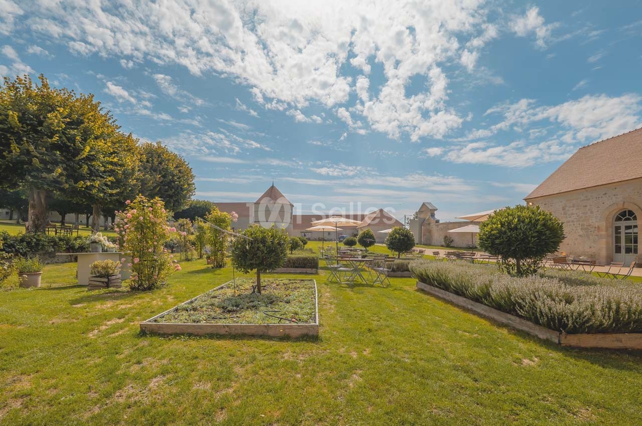 Jardin aménagé avec parterres de fleurs, pelouse et bâtiments en pierre sous un ciel bleu parsemé de nuages.