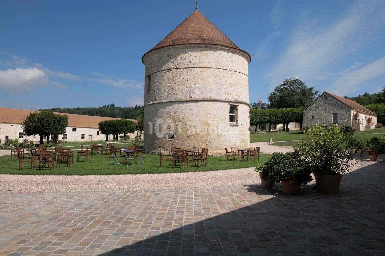 Tour en pierre ronde avec toit conique, entourée de tables en plein air sur une pelouse, dans un cadre rural.