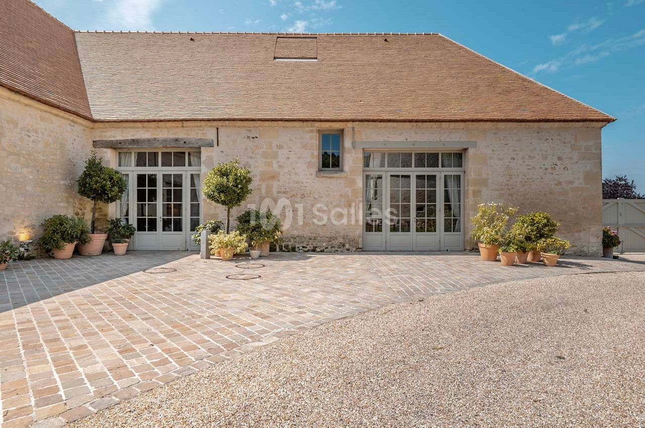Façade d'un bâtiment en pierre avec grandes portes vitrées, entouré de plantes en pots sur une cour pavée.