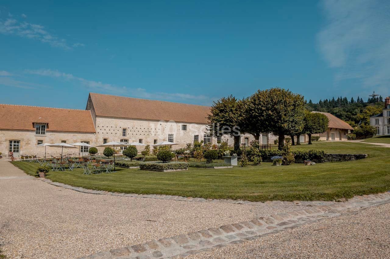 Bâtiment en pierre avec toit en tuiles entouré d'un jardin aménagé, arbres et mobilier de jardin sous un ciel bleu.