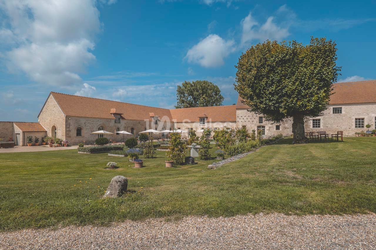 Bâtisse en pierre avec toit en tuiles, entourée d'un jardin verdoyant et d'une terrasse avec parasols.
