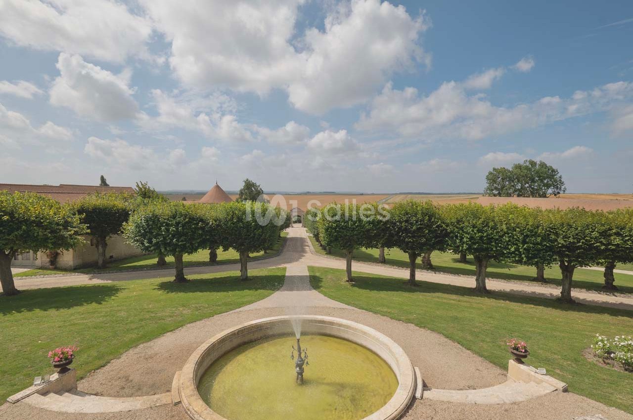 Vue d'une cour avec une fontaine centrale, entourée d'arbres taillés et donnant sur des champs sous un ciel partiellement…