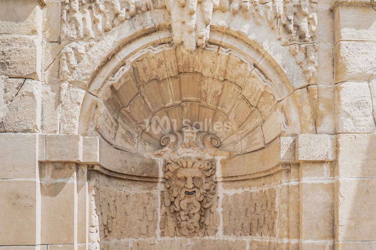 Bas-relief en pierre représentant un visage sculpté sous une arche ornée, intégré à une façade ancienne.