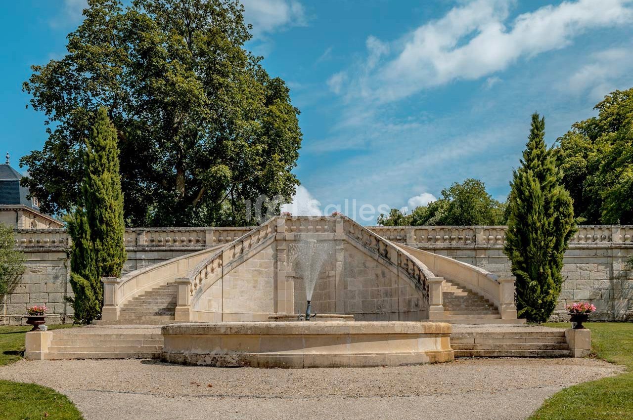 Escalier en pierre symétrique entouré d'arbres, avec une fontaine centrale et un ciel bleu en arrière-plan.