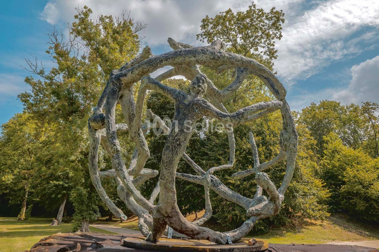 Sculpture en métal représentant une sphère formée de branches entrelacées, exposée dans un parc verdoyant.