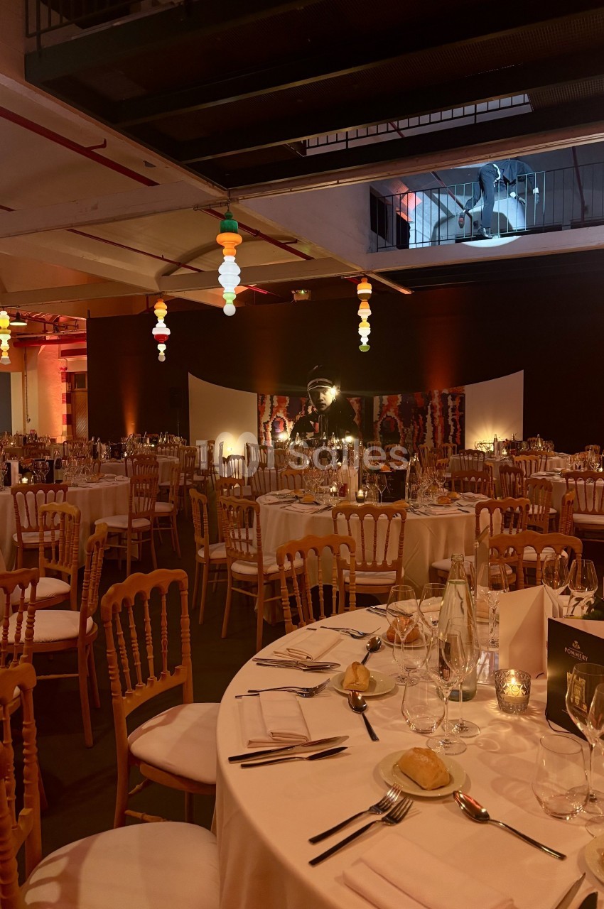 Salle de réception décorée avec des tables rondes dressées, chaises en bois et éclairage chaleureux.