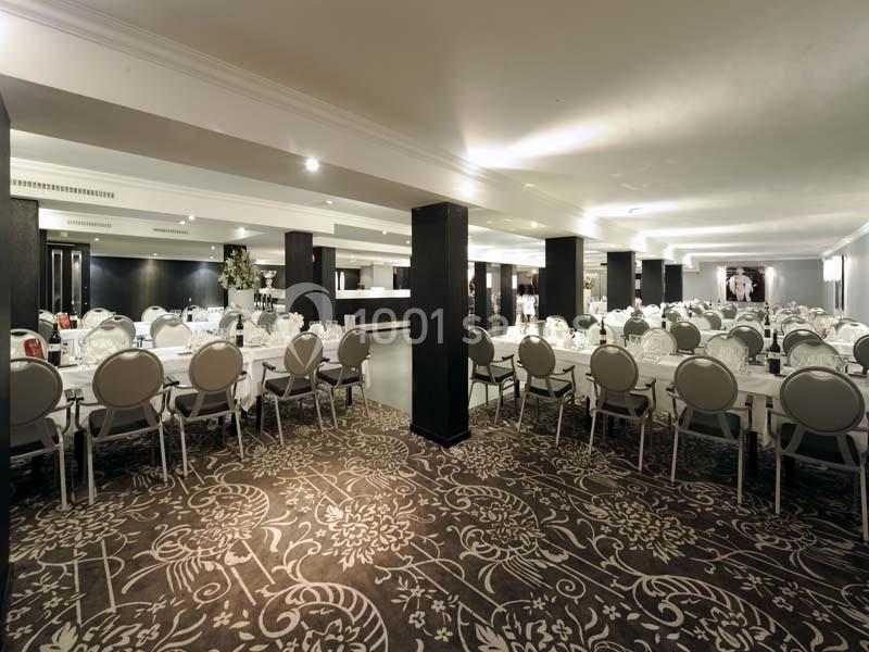 Salle de réception spacieuse avec tables rondes dressées, chaises élégantes et éclairage tamisé.