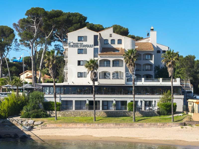 Façade d'un hôtel en bord de mer, entouré de palmiers et de végétation, avec une plage au premier plan.