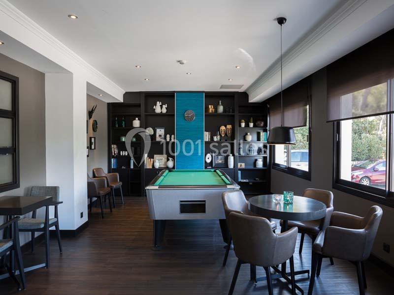 Salle moderne avec un billard central, des tables et des chaises, entourée d'étagères décoratives et de grandes fenêtres.