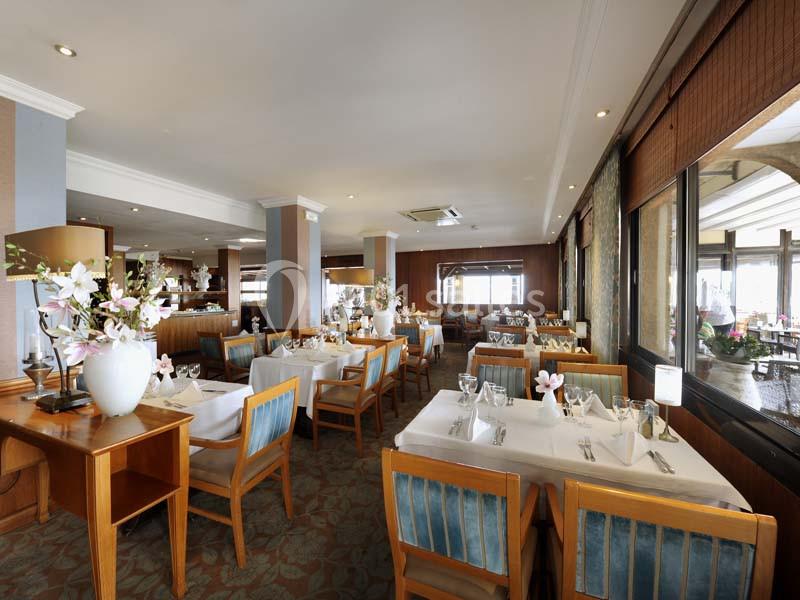 Salle de restaurant lumineuse avec tables dressées, chaises en bois et grandes baies vitrées donnant sur l'extérieur.