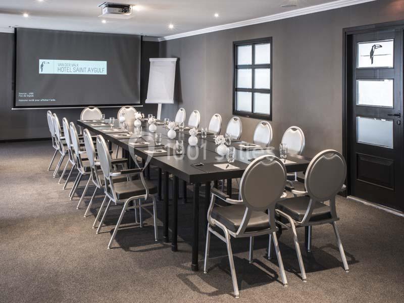 Salle de réunion moderne avec une grande table rectangulaire, des chaises blanches et un écran de projection allumé.