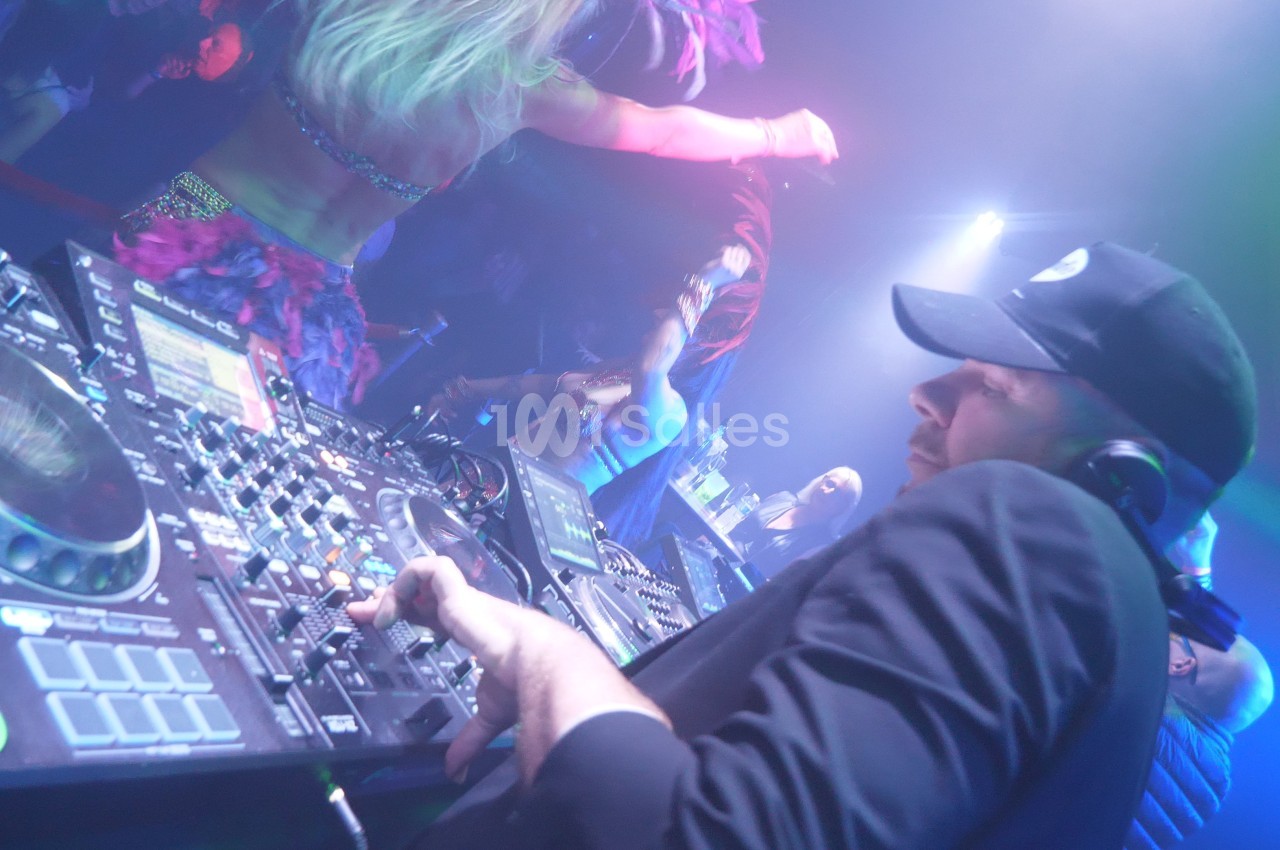 Un DJ mixe sur une console dans une ambiance de fête avec des danseurs et des lumières colorées en arrière-plan.