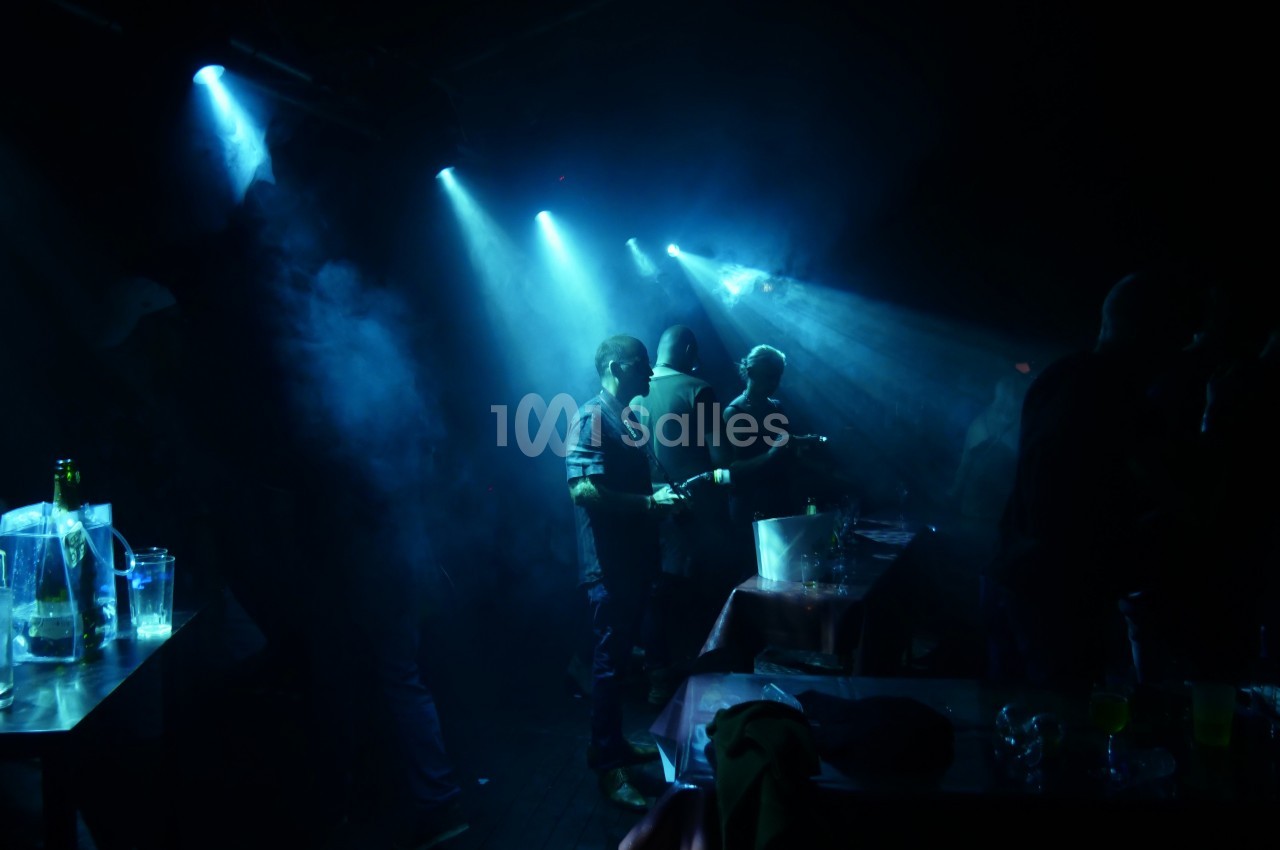 Personnes debout dans une ambiance sombre de club, éclairées par des projecteurs bleus et entourées de fumée.