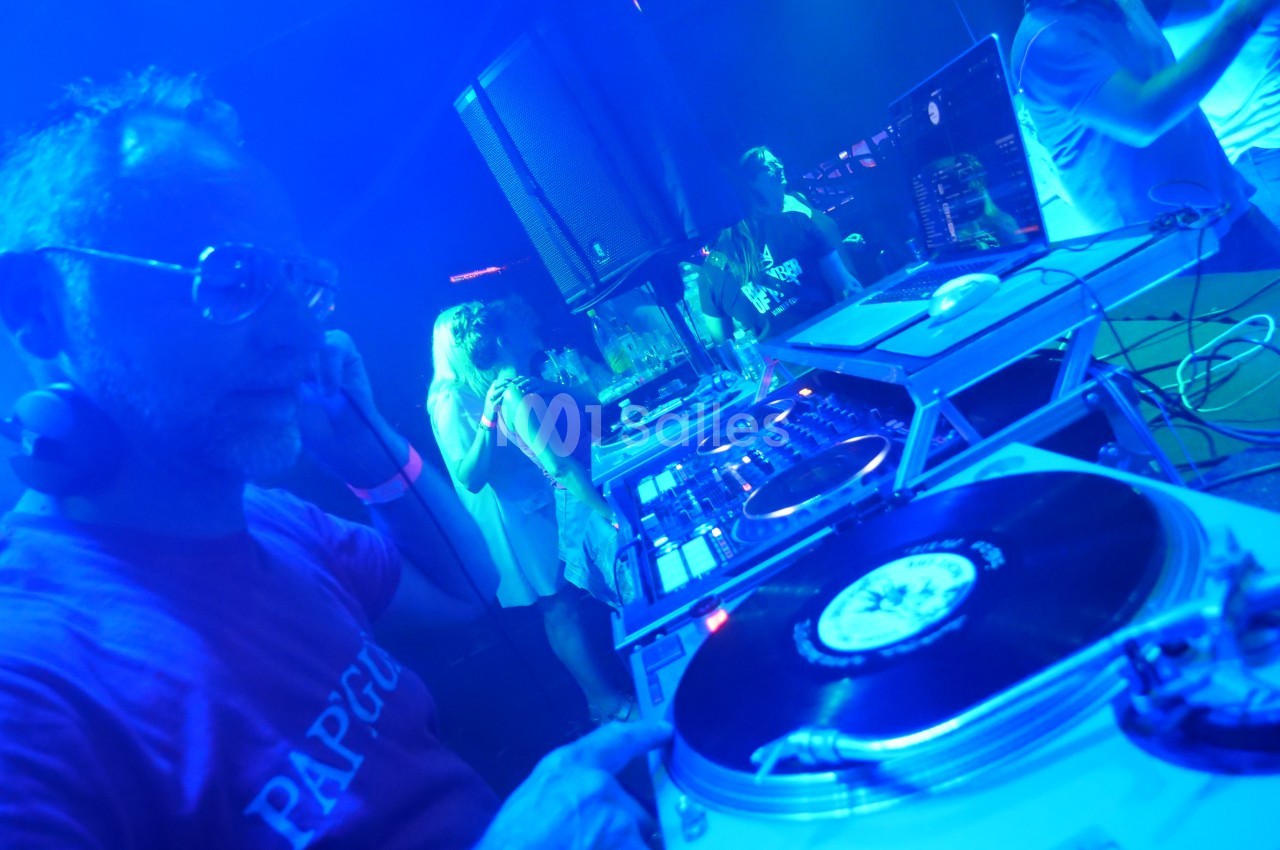 Un DJ mixe des vinyles sur une platine dans une ambiance lumineuse bleutée, entouré de personnes dansant.