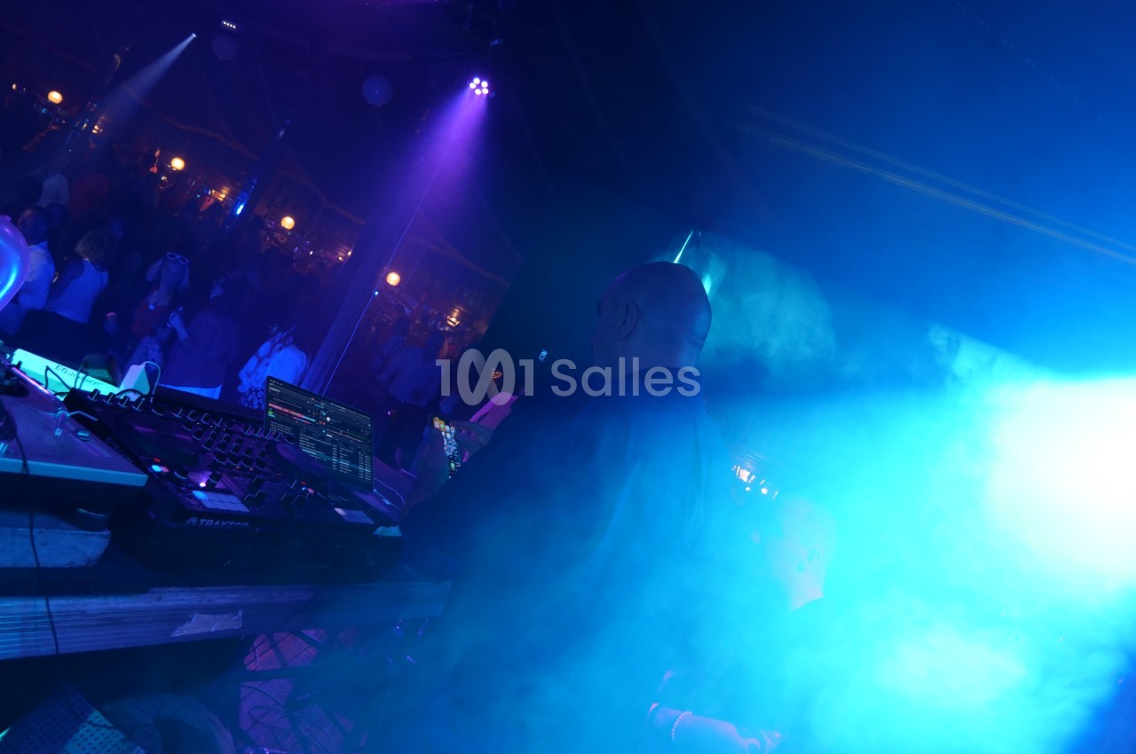 Un DJ mixe de la musique sur scène dans une ambiance lumineuse bleutée, face à une foule dans l'obscurité.