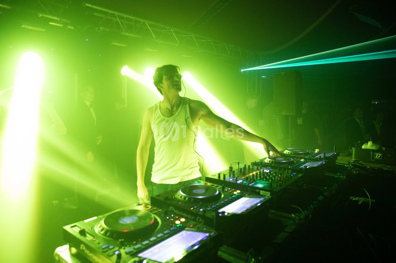 Un DJ en débardeur blanc mixe sur une table de mixage dans une ambiance lumineuse verte et sombre.