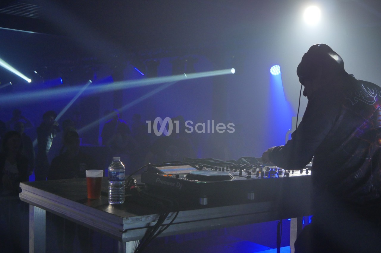 Un DJ mixe sur scène dans une ambiance sombre avec des éclairages bleus et un public en arrière-plan.