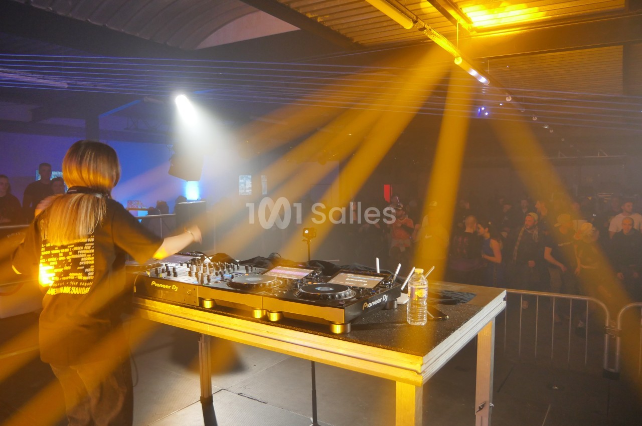 Une DJ mixe sur une scène éclairée par des projecteurs jaunes, face à un public dans une salle sombre.