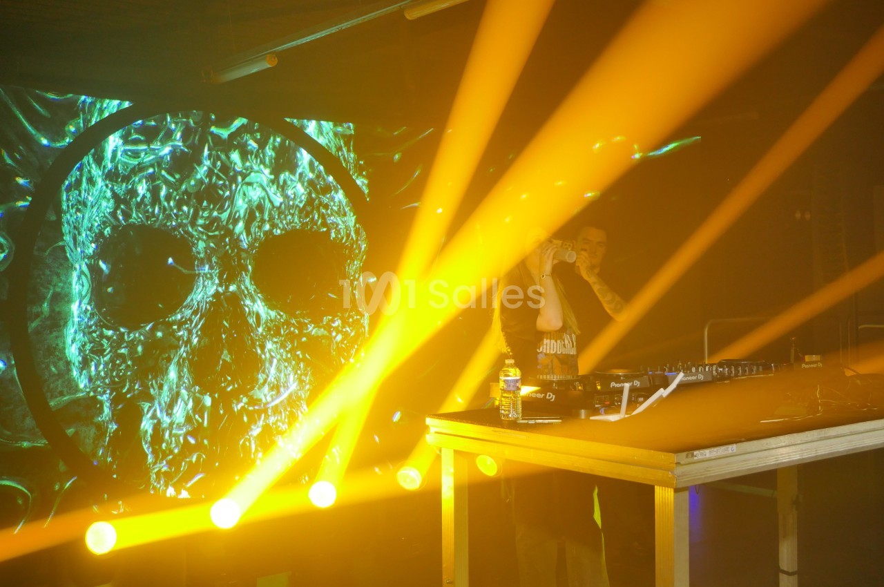 Un DJ mixe sur scène avec des jeux de lumière jaunes et un écran affichant une image de crâne en arrière-plan.